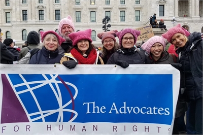 [ai] A group of seven women wearing pink hats poses in front of a banner that reads 'The Advocates for Human Rights' at a public gathering. The background features a historic building and attendees carrying signs.